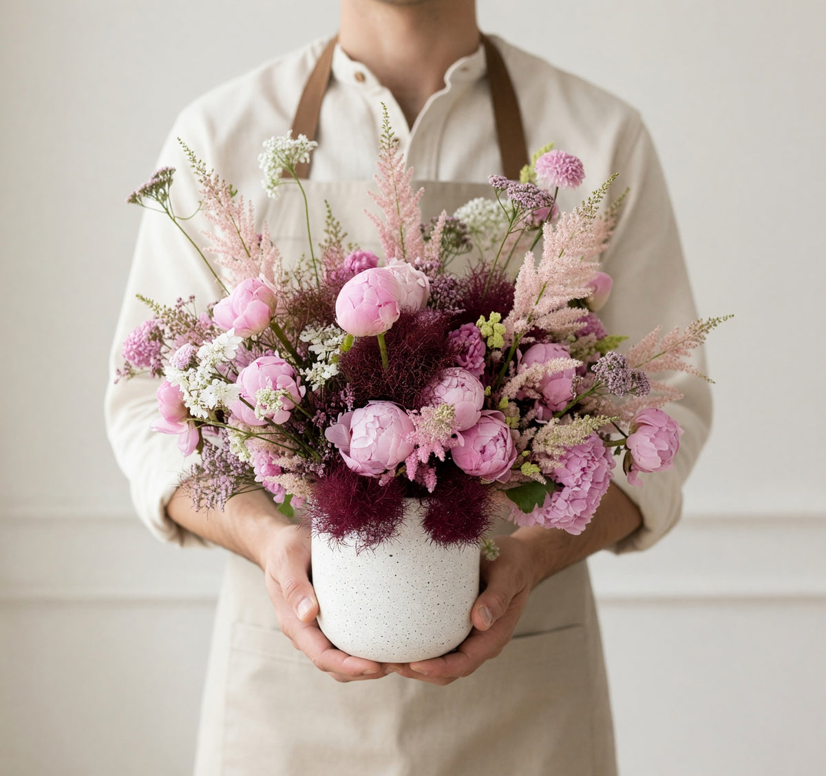 A person wearing a cream shirt and apron holds a white speckled ceramic vase filled with a pink and purple floral arrangement against a light neutral wall.