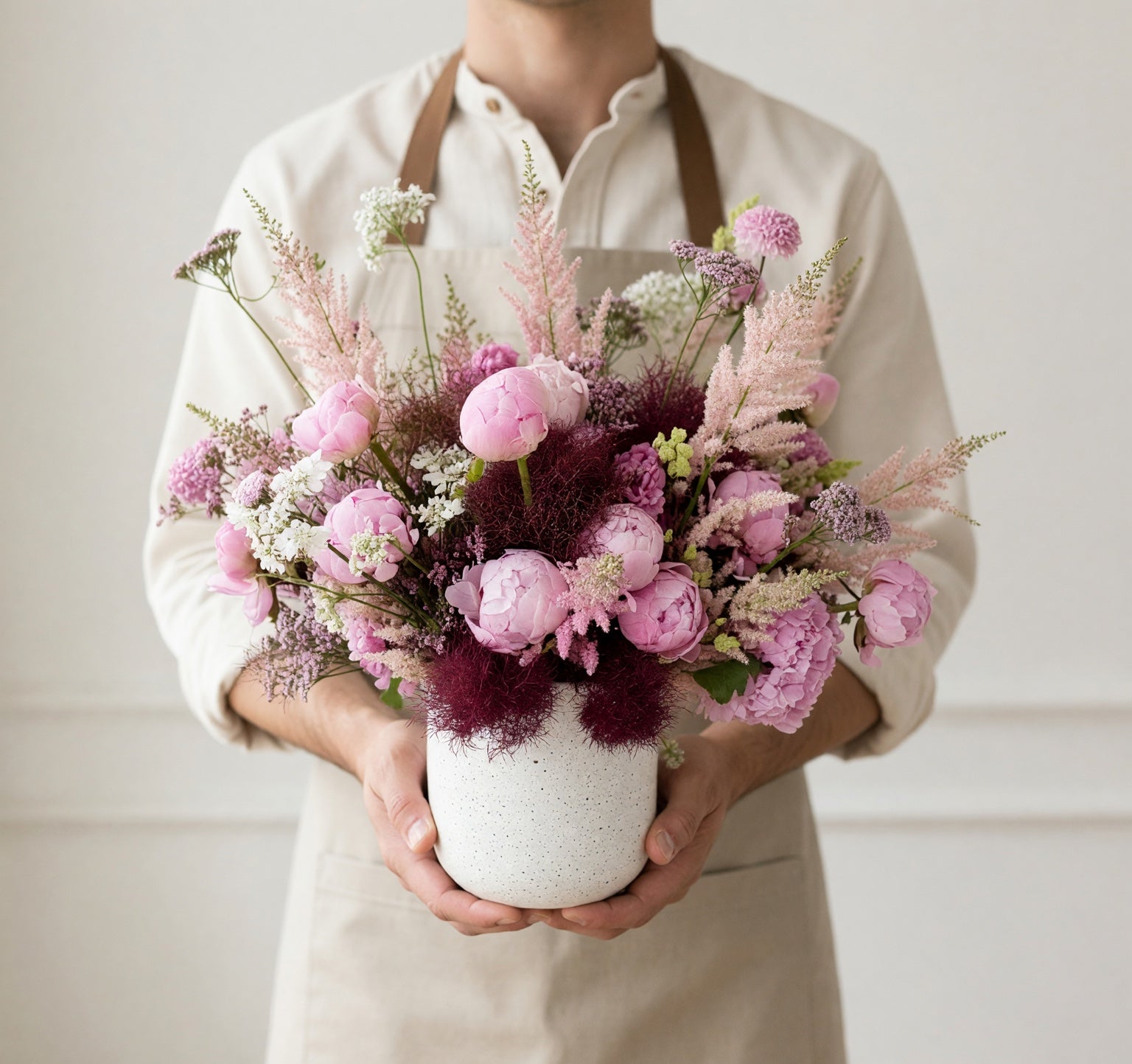A person wearing a cream shirt and apron holds a white speckled ceramic vase filled with a pink and purple floral arrangement against a light neutral wall.