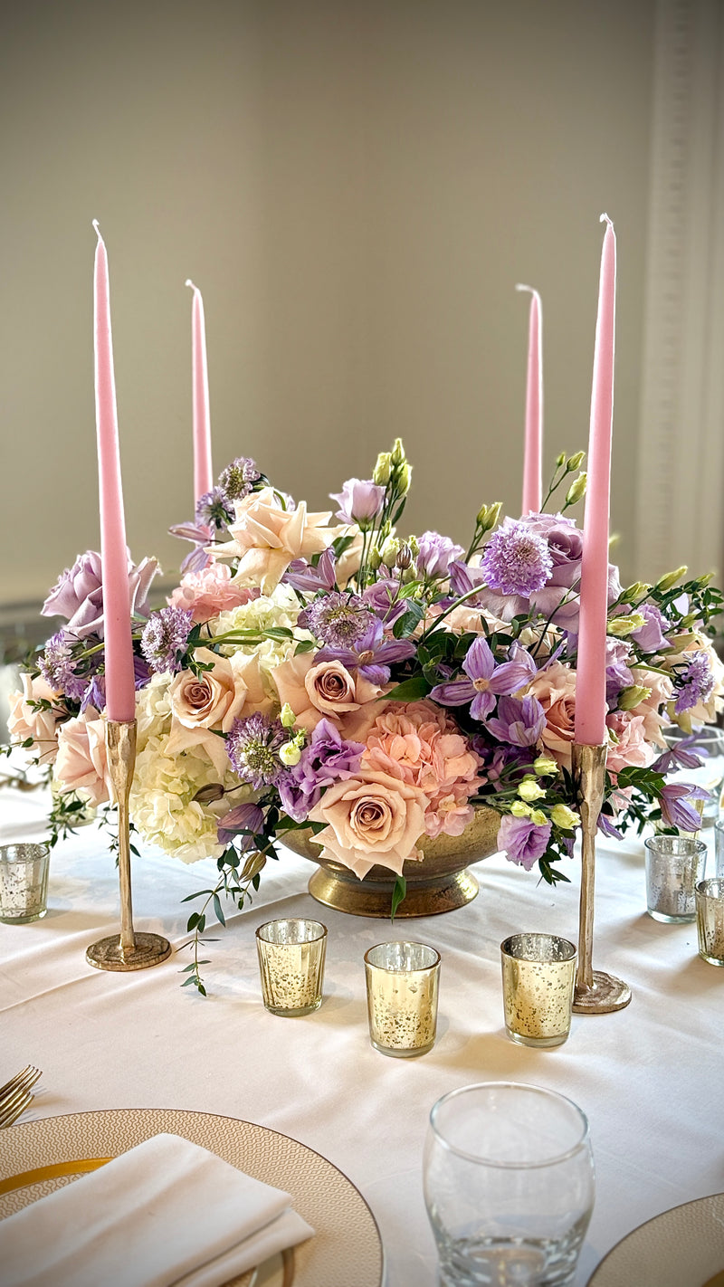 A wedding-style floral centerpiece arrangement in a gold bowl on a white tablecloth, surrounded by four tall pink taper candles in gold holders and small gold votive holders. The bouquet includes blush pink, lavender, cream, and purple flowers with green foliage.