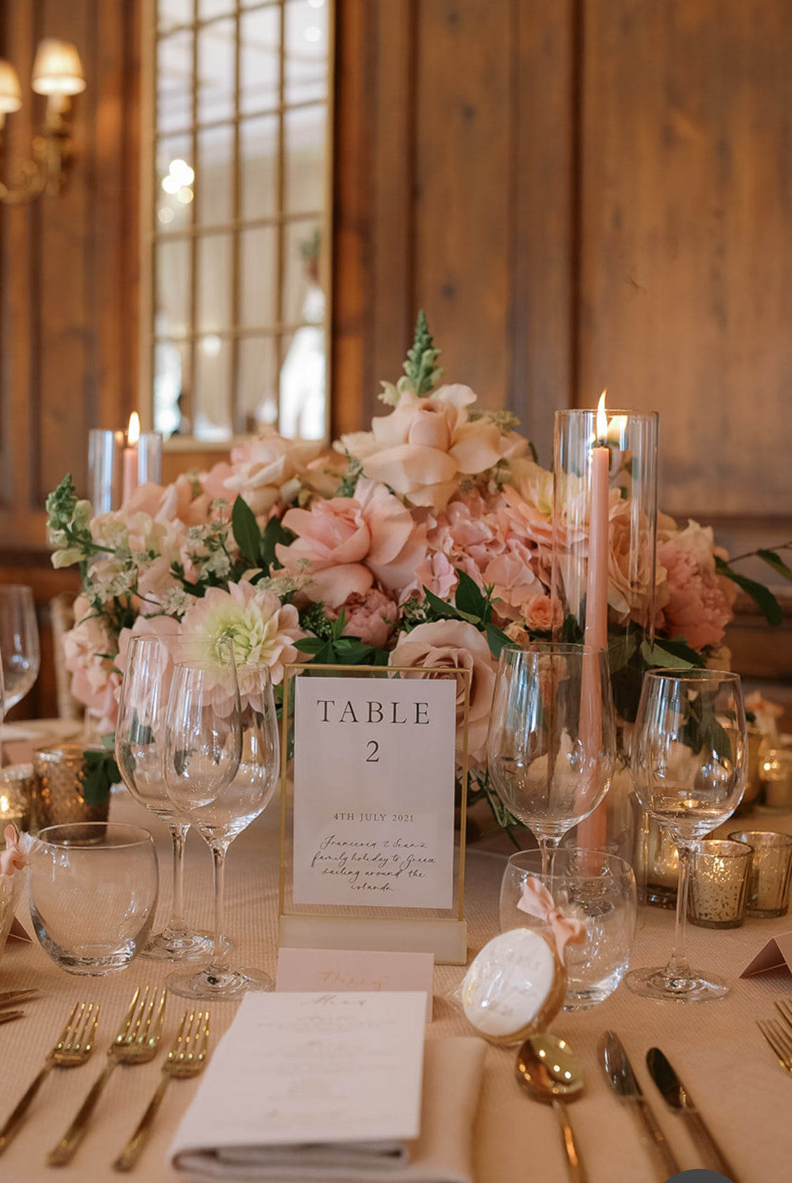 A styled wedding table centerpiece with a large arrangement of blush pink and white flowers, clear glass candle holders with lit taper candles, wine glasses, gold flatware, and a table place card reading TABLE 2 in a rustic wood-paneled room.
Details