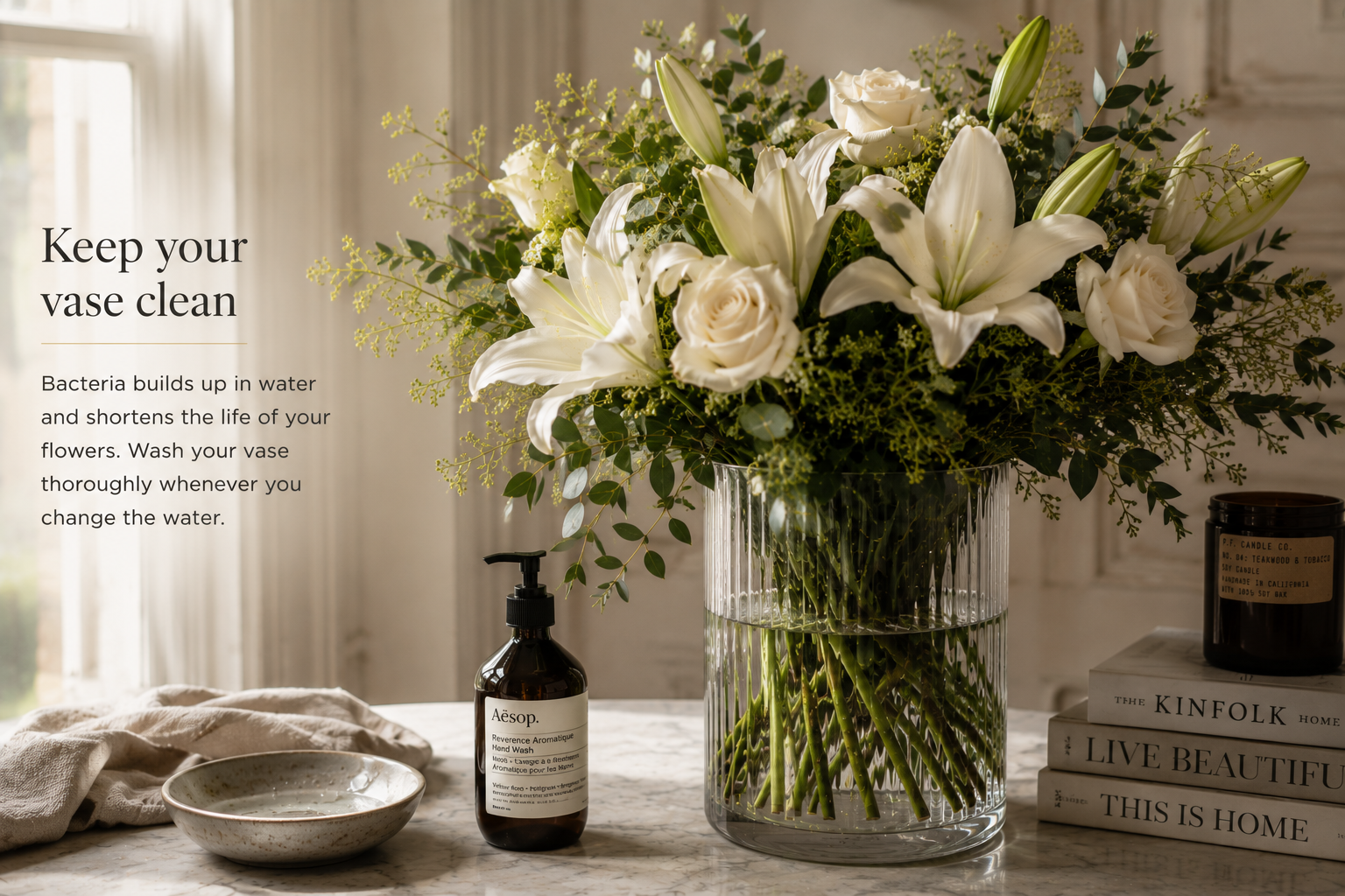 A large floral arrangement in a ribbed clear glass vase sits on a tabletop beside a small amber pump bottle, a folded cloth, and books in a softly lit interior. Text on the left reads, 'Keep your vase clean' with a brief explanatory note about bacteria in water and flower longevity.