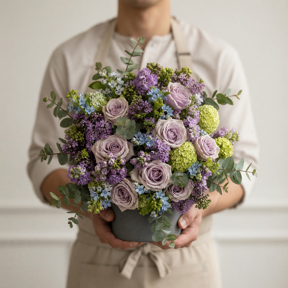 A person in a beige shirt and apron holds a large, round floral arrangement in a gray pot. The bouquet features lavender-purple roses, light green hydrangeas, blue filler flowers, and green eucalyptus leaves.