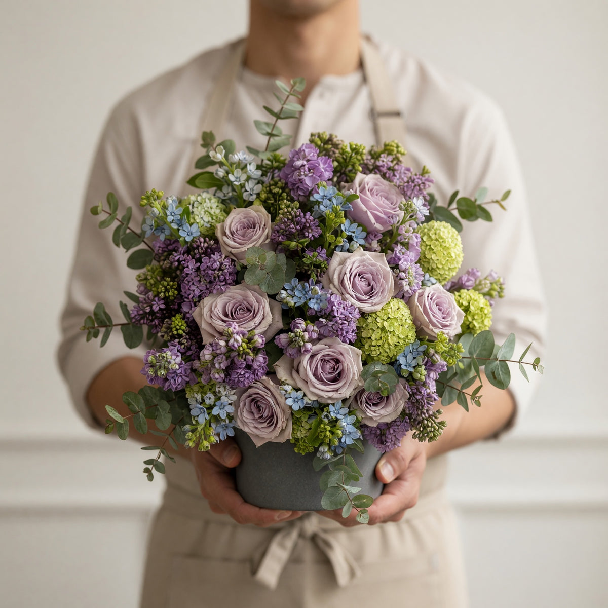 A person in a beige shirt and apron holds a large, round floral arrangement in a gray pot. The bouquet features lavender-purple roses, light green hydrangeas, blue filler flowers, and green eucalyptus leaves.