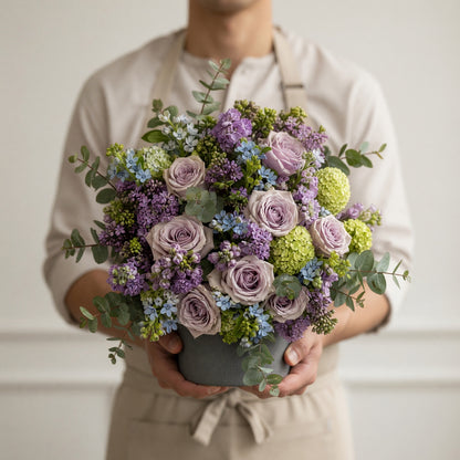 A person in a beige shirt and apron holds a large, round floral arrangement in a gray pot. The bouquet features lavender-purple roses, light green hydrangeas, blue filler flowers, and green eucalyptus leaves.